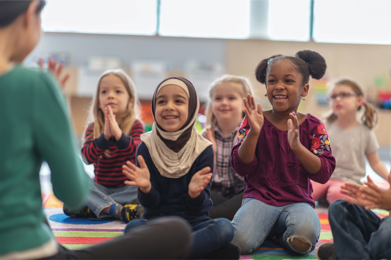 A group of kids are clapping along to music. They are sitting in their classroom.
